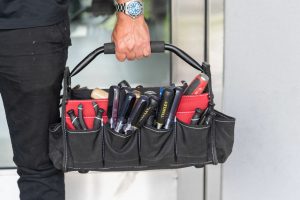 A person holds a black tool bag filled with various hand tools, including screwdrivers and a Stanley utility knife.