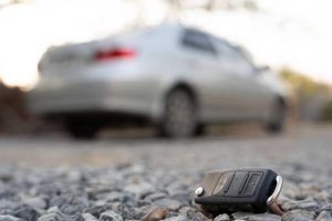 Car key fob on gravel with a blurred silver sedan in the background.
