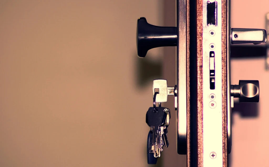 A close-up of a door edge showing a lock with a set of keys inserted, set against a blurred beige background.