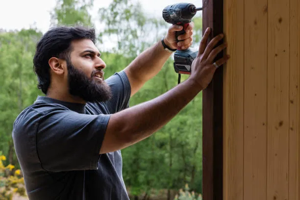 A person uses a cordless power drill to fasten or secure a piece of wood to a vertical wooden structure outdoors.