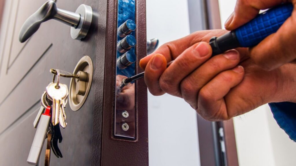 A person uses tools to pick a door lock while a set of keys with keychains hangs from the lock.