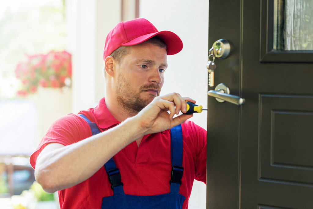 A locksmith in a red cap and blue overalls uses a screwdriver to repair a door lock.